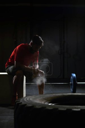 Photo for Stock photo of an adult man in a gym sitting down and resting. There is a tractor tire in front and a barbell behind him. He is wearing sportswear. - Royalty Free Image