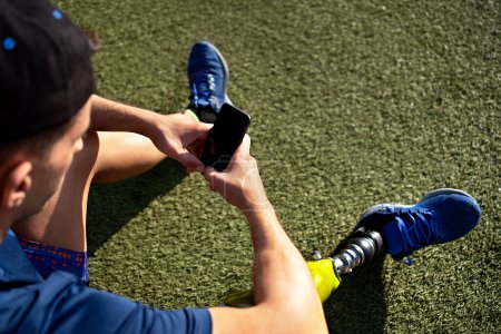 Téléchargez les photos : Athlète homme handicapé utilisant son téléphone tout en étant assis dans l'herbe. - en image libre de droit