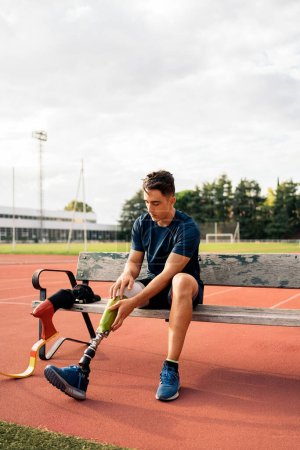 Photo for Stock photo of disabled man athlete sitting in a bench and putting on his leg prosthesis. - Royalty Free Image