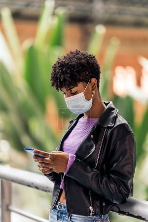 Photo for Stock photo of african girl wearing face mask using phone while waiting in train station. - Royalty Free Image