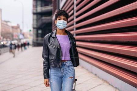 Photo for Stock photo of young african girl wearing face mask and carrying suitcase walking in the city. - Royalty Free Image