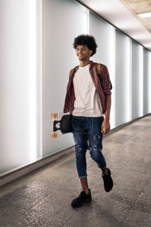 Photo for Stock photo of young afro boy walking in the city and carrying his longboard. - Royalty Free Image