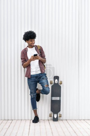 Photo for Stock photo of cool african boy standing next to his longboard smiling and using cellphone. - Royalty Free Image