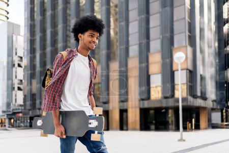 Téléchargez les photos : Photo de stock de jeune afro garçon marchant dans la ville portant son longboard. - en image libre de droit