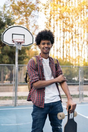 Téléchargez les photos : Photo de stock de beau jeune homme africain avec longboard souriant et regardant à la caméra dans le terrain de basket-ball. - en image libre de droit