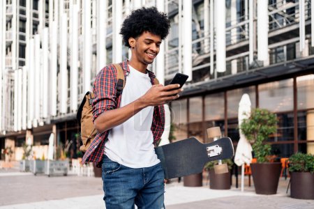 Téléchargez les photos : Photo de stock de jeune afro garçon marchant dans la ville et portant son longboard. - en image libre de droit