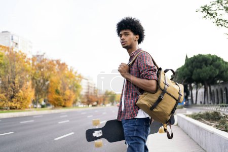 Téléchargez les photos : Photo de stock de jeune afro garçon marchant dans la ville et portant son longboard. - en image libre de droit