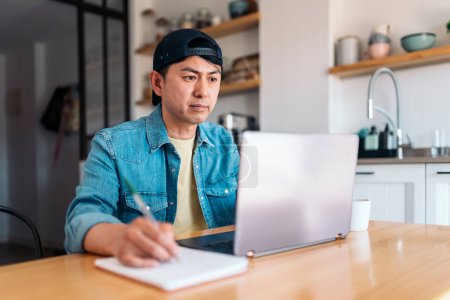 Téléchargez les photos : Asiatique homme écriture dans notebook tout en utilisant ordinateur portable dans la cuisine - en image libre de droit