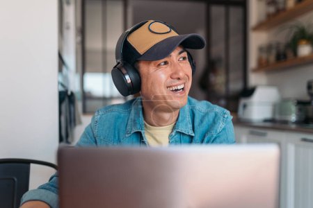 Téléchargez les photos : Heureux asiatique casual homme d'affaires à l'aide d'écouteurs lors d'une réunion en ligne tout en travaillant à la maison - en image libre de droit
