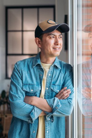 Photo for Vertical portrait of a relaxed asian man looking away through the window standing at home - Royalty Free Image