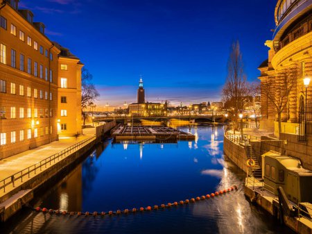 Foto de Estocolmo, Suecia - 6 de febrero de 2023: Un paisaje urbano de monumentos iluminados que se reflejan en la vía navegable de color azul en el crepúsculo, proporcionando una impresionante vista nocturna hacia el Ayuntamiento de Estocolmo - Imagen libre de derechos