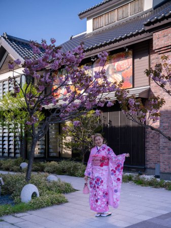 A woman in a traditional pink kimono poses happily under blooming cherry blossoms in front of traditional Japanese architecture