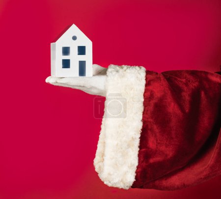 Hand in Hand in roter Weihnachtsmann-Tracht hält ein Modell eines weißen Hauses. Geschäft von Immobilien, Wohnimmobilien zu verkaufen und zu vermieten - Kopierraum, Werbedesign inserieren.