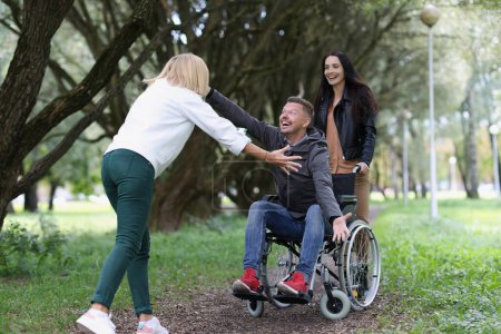 Téléchargez les photos : Portrait de femmes amis marchant avec un homme handicapé en fauteuil roulant dans le parc, heureux de se voir. Invalidité et concept de santé - en image libre de droit