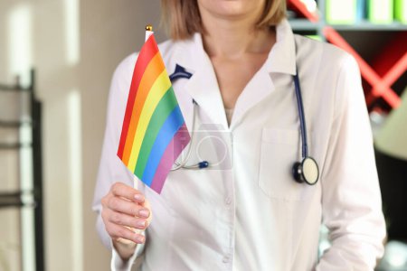 Téléchargez les photos : Woman doctor shows lgbt pride flag in her hand in hospital office. Medical support for lgbt community and symbol of tolerance. - en image libre de droit