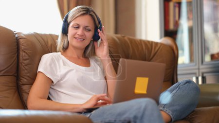 Photo for Woman with headphones smiling and looking at laptop screen while sitting on the couch. Freelance manager speaking with client online at home. - Royalty Free Image