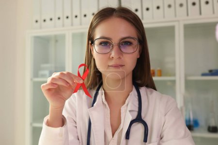 Téléchargez les photos : Jeune femme médecin dans des lunettes tenant un ruban rouge comme symbole de la maladie du sida. Journée mondiale du sida, sensibilisation au VIH. - en image libre de droit