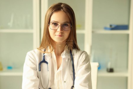 Téléchargez les photos : Jeune jolie docteur en lunettes avec stéthoscope regardant la caméra. Stagiaire féminine travaillant dans une clinique médicale, concept d'éducation médicale. - en image libre de droit