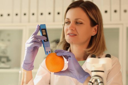 Téléchargez les photos : Une femme dans le laboratoire tient un appareil et une agrumes orange, un gros plan. Vérification des fruits pour les nitrates - en image libre de droit