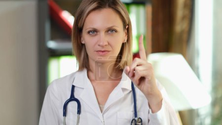 Téléchargez les photos : Femme médecin avec stéthoscope montre geste avec son index vers le haut. Symbole d'évitement, recommandations et rappels. - en image libre de droit