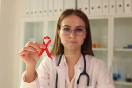 Téléchargez les photos : Une jeune virologiste portant des lunettes tient un ruban rouge comme symbole de la maladie du sida à l'hôpital. Journée internationale du sida, sensibilisation au VIH. - en image libre de droit