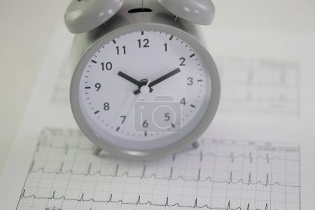 Foto de Reloj despertador redondo en papeles de cardiograma en la mesa del hospital. Monitoreo profesional de frecuencia cardíaca. Primer plano del concepto de atención sanitaria y esperanza de vida - Imagen libre de derechos