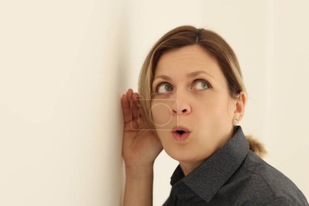 Curious woman listens to discussion of colleagues in office. Attentive female person with surprised facial expression puts ear close to wall