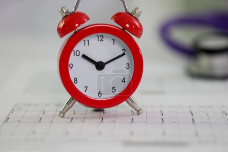 Foto de Reloj despertador rojo en papel cardiograma en la mesa de la clínica. Monitorización profesional de la frecuencia cardíaca en el hospital. Concepto de salud y esperanza de vida - Imagen libre de derechos