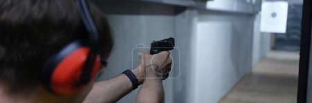 Téléchargez les photos : Close-up of guy in tactical headphones aiming pistol at target in shooting range. Shooting practicing with gun in rifle range. Extreme hobby concept - en image libre de droit