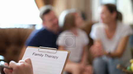 Psychologist ready to make notes listening to complaints of family. Specialist holds clipboard with blank paper against parents and kid on sofa
