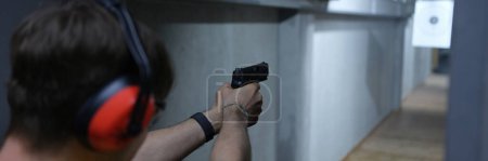 Téléchargez les photos : Close-up of man shooter in tactical headphones aiming pistol at target in shooting range. Shooting practicing with gun in rifle range. Extreme hobby concept - en image libre de droit