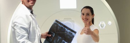 Téléchargez les photos : Radiologist holds MRI scans results while standing near CT scanner and patient. Female patient shows thumbs up gesture after medical diagnostic. - en image libre de droit
