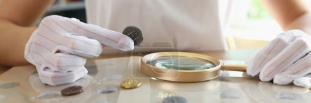 Photo for Numismatist in white gloves examines ancient coins collection at table. Close-up of woman with magnifying glass values coins. - Royalty Free Image