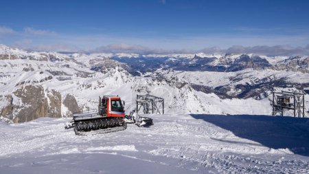 This image features a breathtaking view of a snow-covered mountain range under a clear blue sky. A red snowcat, designed for snowy terrains, is prominently featured in the foreground.