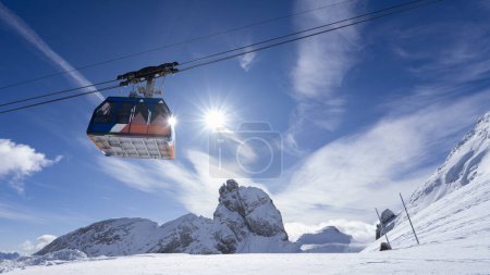 Image captures a snowy landscape under a bright blue sky. A colorful cable car is suspended in mid-air, traveling across the scene. The sun illuminates the snowy peaks, creating a serene atmosphere