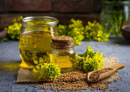Photo for Mustard oil in a glass jar and decanter, mustard grains on a burlap napkin, flowers and leaves on wooden board background - Royalty Free Image