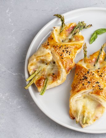 Téléchargez les photos : Baked green asparagus with ham and cheese in puff pastry sprinkled with sesame seeds and green basil leaves. - en image libre de droit