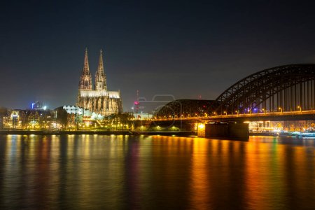 Foto de Vista panorámica del puente iluminado sobre el río Rin donde pasan trenes y turistas y la catedral de Colonia por la noche - Imagen libre de derechos