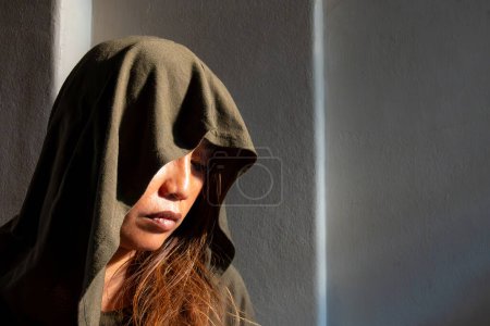 A solitary woman in a hood gazes downward, her face illuminated by a stark contrast of natural sunlight and shadow, evoking a thoughtful mood.