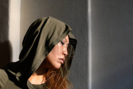 Una mujer en una capucha es capturada en un rayo de luz solar sobre un fondo sombrío, creando un contraste llamativo que resalta su estado de ánimo contemplativo..