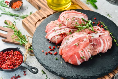 Photo for Raw cutlets wrapped in bacon served on a black plate with rosemary and thyme. On a stone background. Top view. - Royalty Free Image