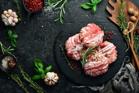 Photo for Pork cutlets wrapped in bacon served on a black plate with rosemary and thyme. On a black stone background. Top view. - Royalty Free Image