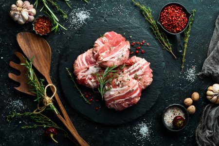 Photo for Pork cutlets wrapped in bacon served on a black plate with rosemary and thyme. On a black stone background. Top view. - Royalty Free Image