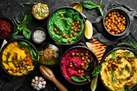 Photo for Colorful hummus bowls - green, yellow and beetroot hummus on dark background with lemon, olive oil, and spices. On a concrete black background. - Royalty Free Image