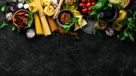 Téléchargez les photos : Spaghettis secs et ingrédients pour leur préparation : basilic, parmesan, pignons, tomates, épices. Cuisine traditionnelle italienne. Sur un fond de pierre noire. - en image libre de droit