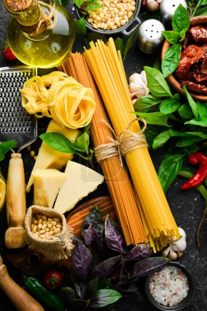 Téléchargez les photos : Spaghettis secs et ingrédients pour leur préparation : basilic, parmesan, pignons, tomates, épices. Cuisine traditionnelle italienne. Sur un fond de pierre noire. - en image libre de droit