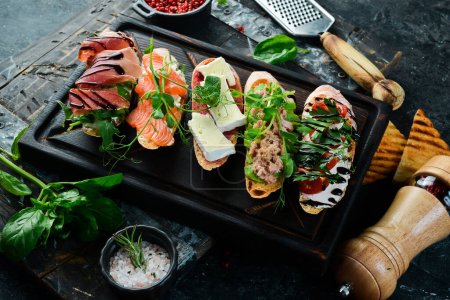 Photo for Sandwich Set bruschetta with tuna, cheese, prosciutto and tomatoes on a dark wooden board. On a black stone background. - Royalty Free Image