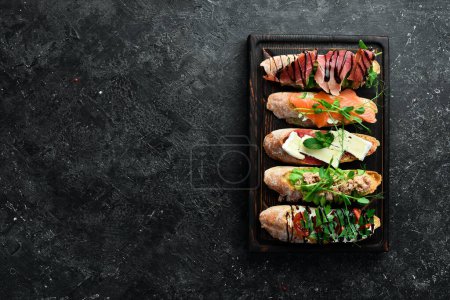 Foto de Sandwich Conjunto de bruschetta con atún, queso, jamón y tomate sobre una tabla de madera oscura. Sobre un fondo de piedra negra. - Imagen libre de derechos