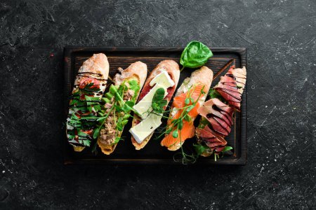 Photo for Sandwich Set bruschetta with tuna, cheese, prosciutto and tomatoes on a dark wooden board. On a black stone background. - Royalty Free Image