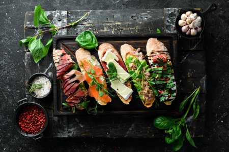 Foto de Sandwich Conjunto de bruschetta con atún, queso, jamón y tomate sobre una tabla de madera oscura. Sobre un fondo de piedra negra. - Imagen libre de derechos